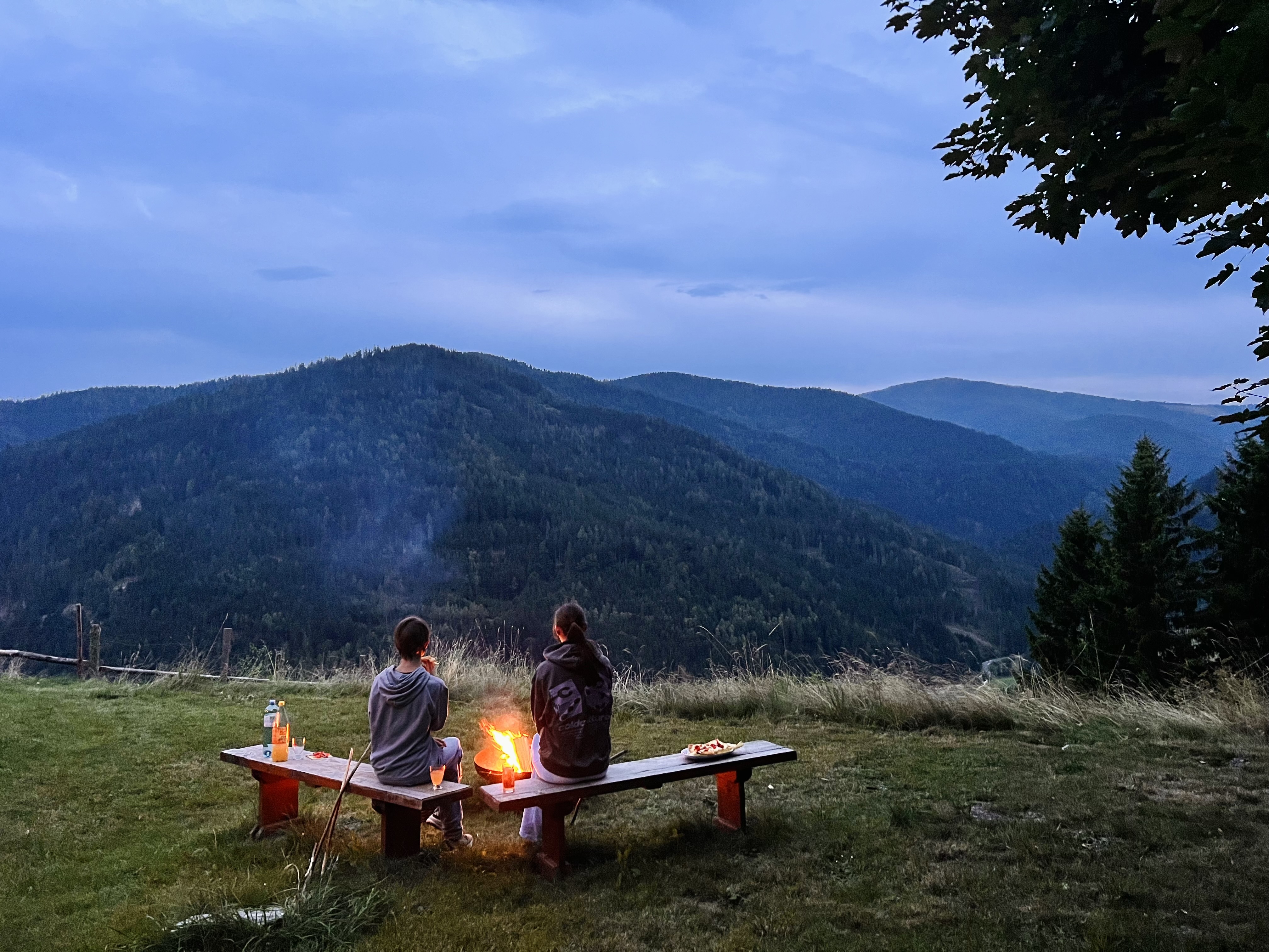 Zwei Personen sitzen auf einer Bank vor der Feuerschale und blicken auf die abendlichen Berge