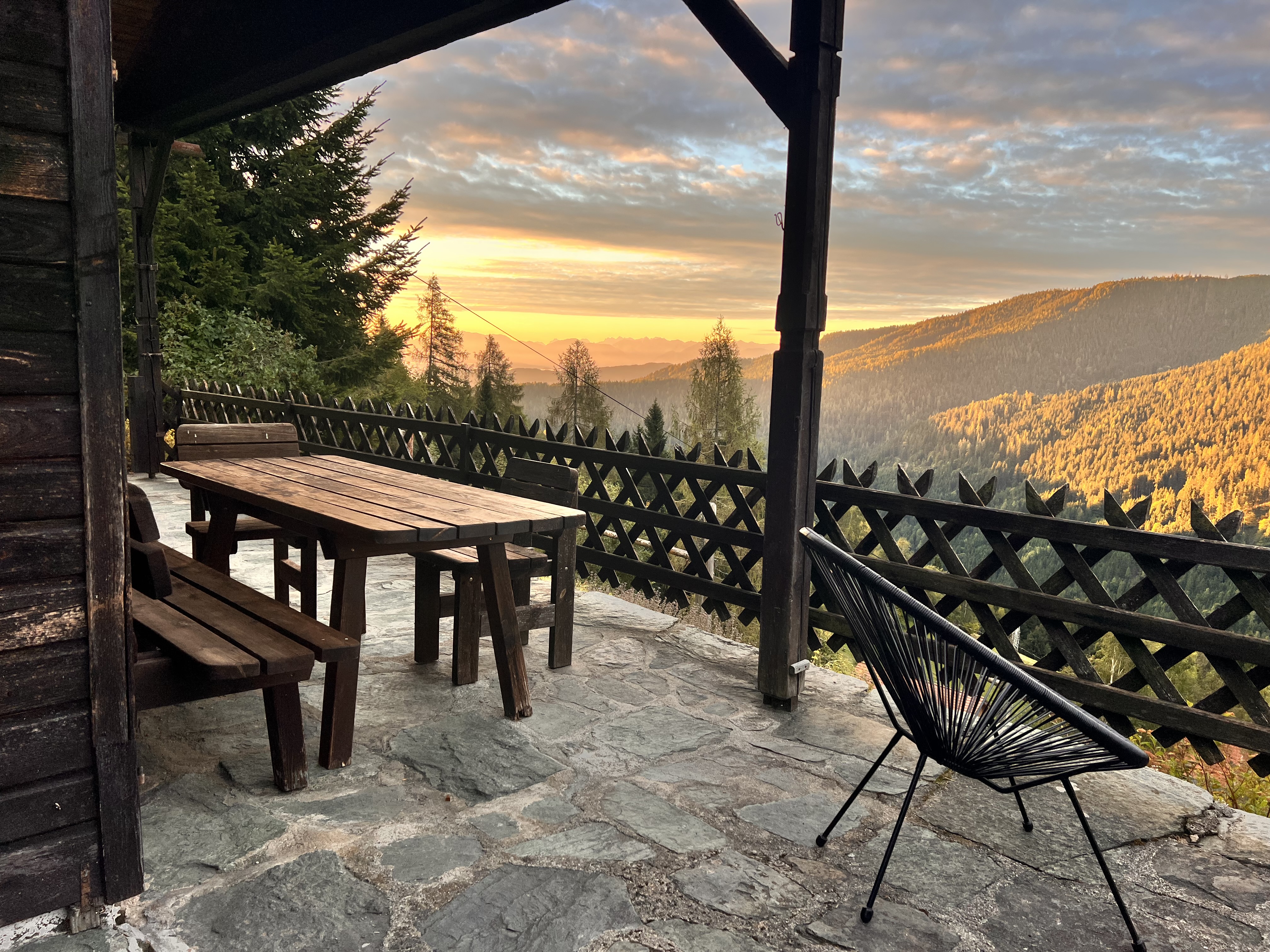 Sonnenuntergang über der Sonnenterrasse der alm44 — Holztisch, Bank, Acapulco-Stuhl im warmen Licht
